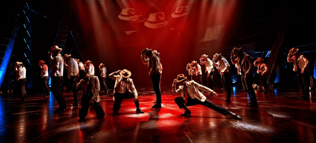 Danseurs en costume clair et chapeau feutre dans une chorégraphie synchronisée sous une lumière rouge, Odyssée
